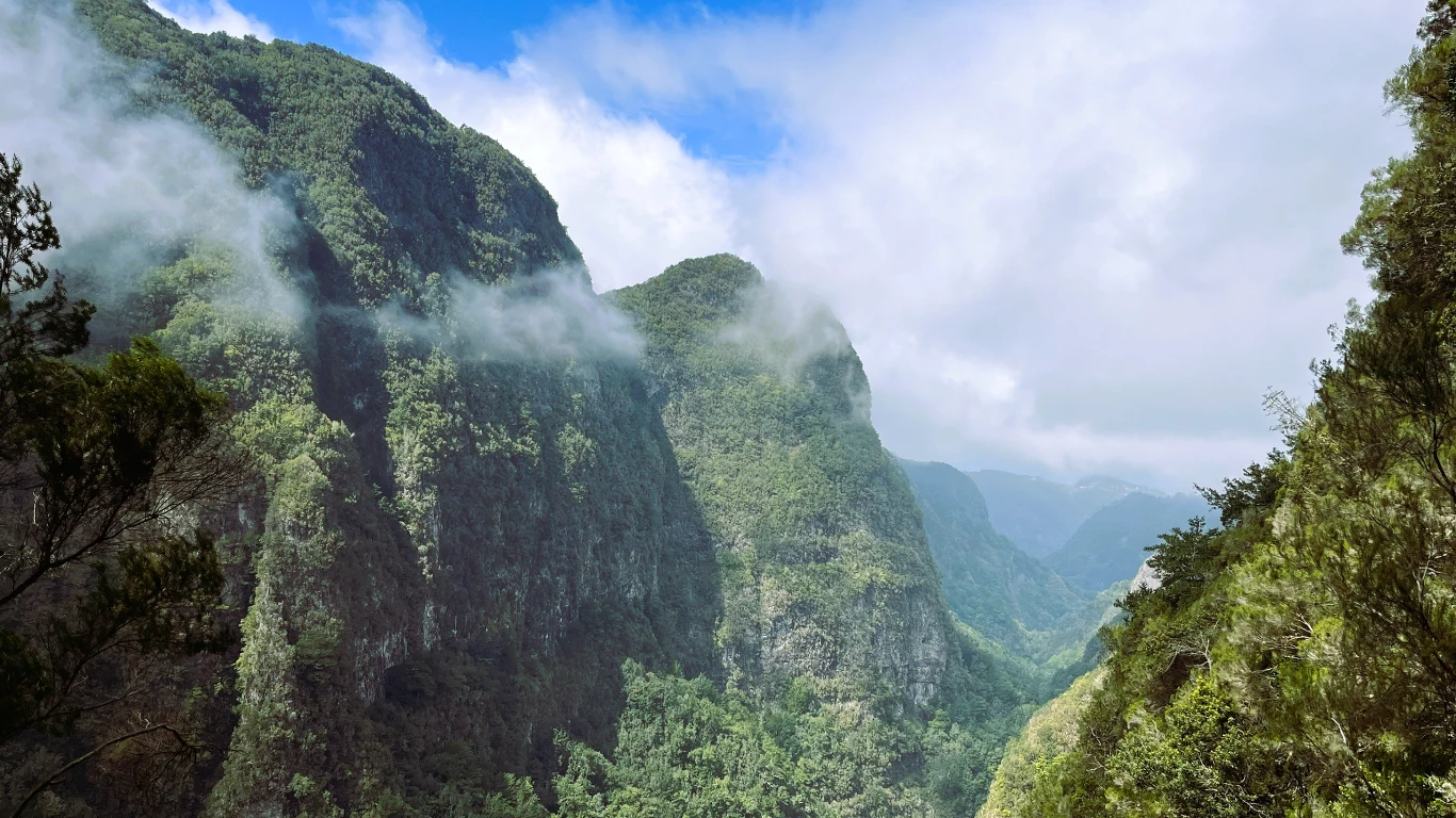 Wysokie, zielone zbocza gór porośnięte bujną roślinnością, częściowo przesłonięte niskimi chmurami i mgłą. W tle widać głęboką dolinę oraz fragment niebieskiego nieba z obłokami.