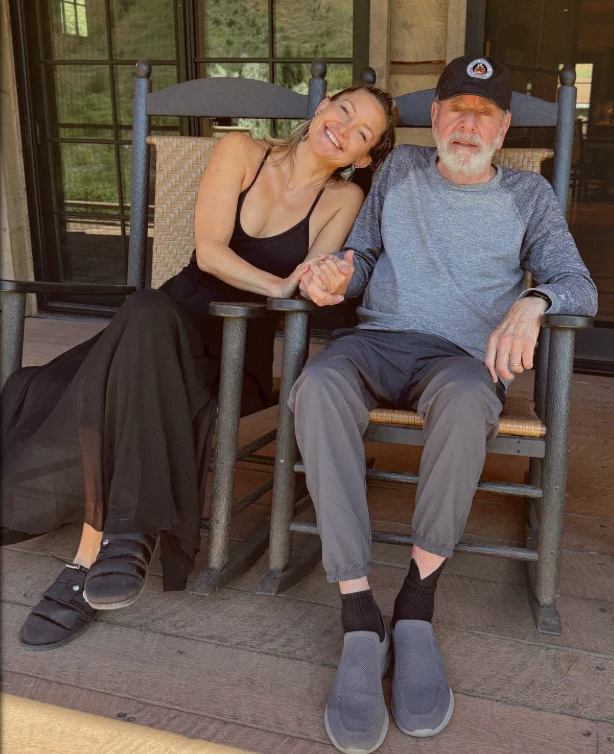 Kobieta w czarnej sukience siedzi na drewnianym bujanym fotelu i opiera głowę na ramieniu starszego mężczyzny w czapce z daszkiem, który uśmiecha się, trzymając ją za rękę. Oboje wyglądają na zrelaksowanych, siedząc na drewnianym tarasie.