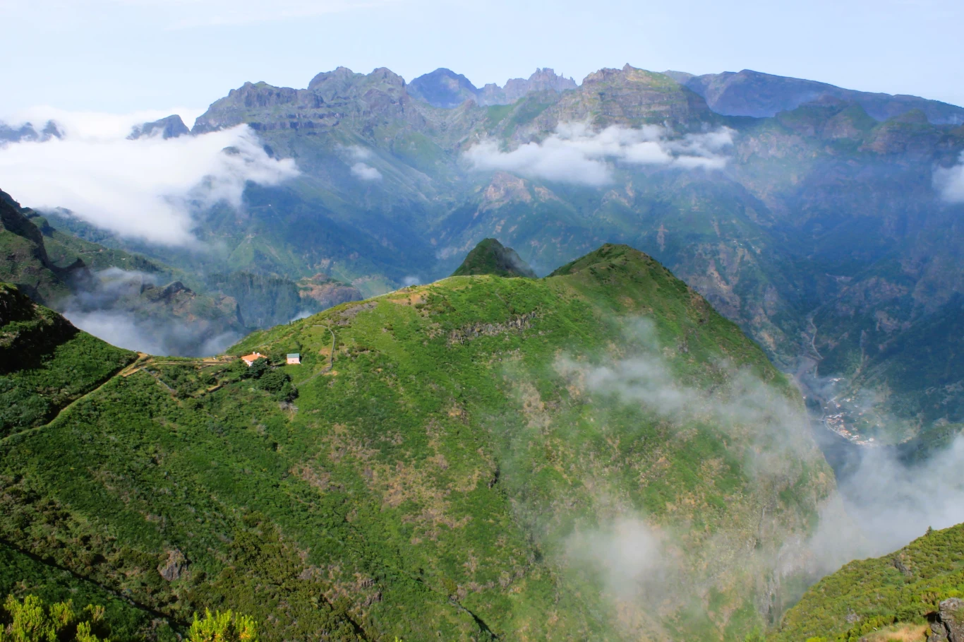 Soczystozielone wzgórza otoczone mgłą i niskimi chmurami, w oddali masywne szczyty górskie oraz fragment małego domu na zboczu. Na pierwszym planie różnorodna roślinność.