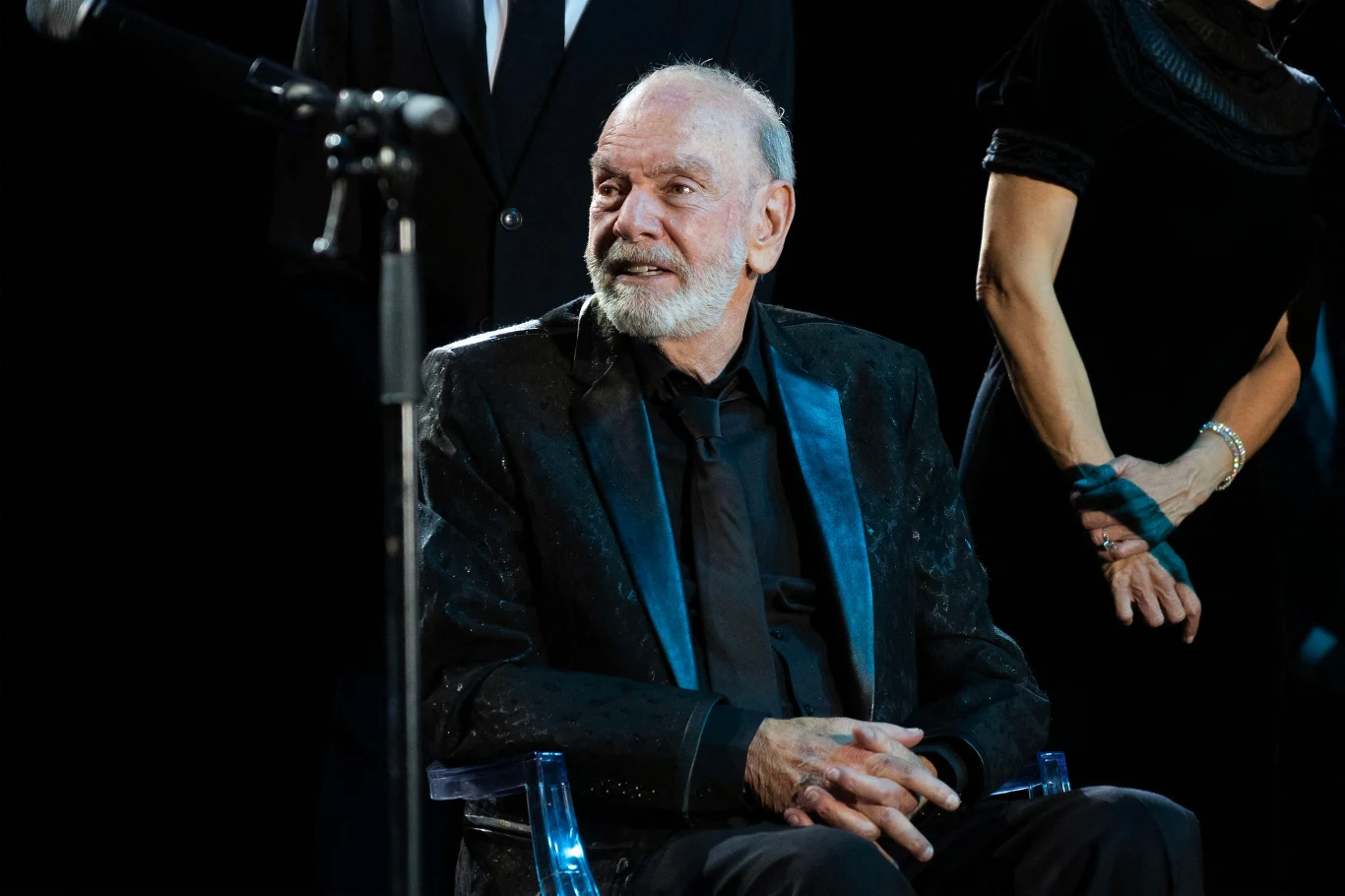 Starszy mężczyzna w czarnym garniturze i krawacie siedzi na krześle, uśmiechając się lekko, obok niego stoją osoby w eleganckich strojach, w tle widoczne jest profesjonalne oświetlenie sceniczne.