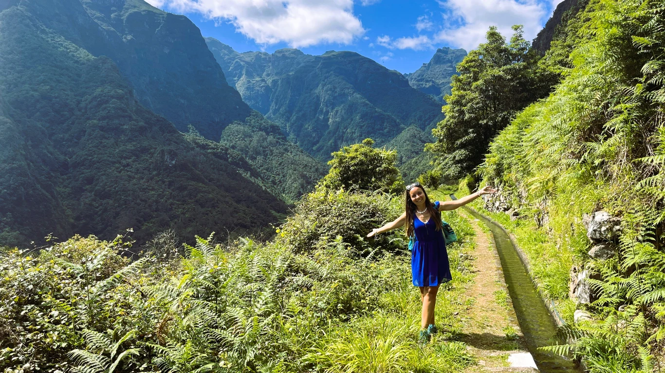 Młoda kobieta w niebieskiej sukience stoi z rozłożonymi ramionami na wąskiej ścieżce otoczonej soczyście zieloną roślinnością, w tle majestatyczne, porośnięte lasem góry pod błękitnym niebem z białymi chmurami.