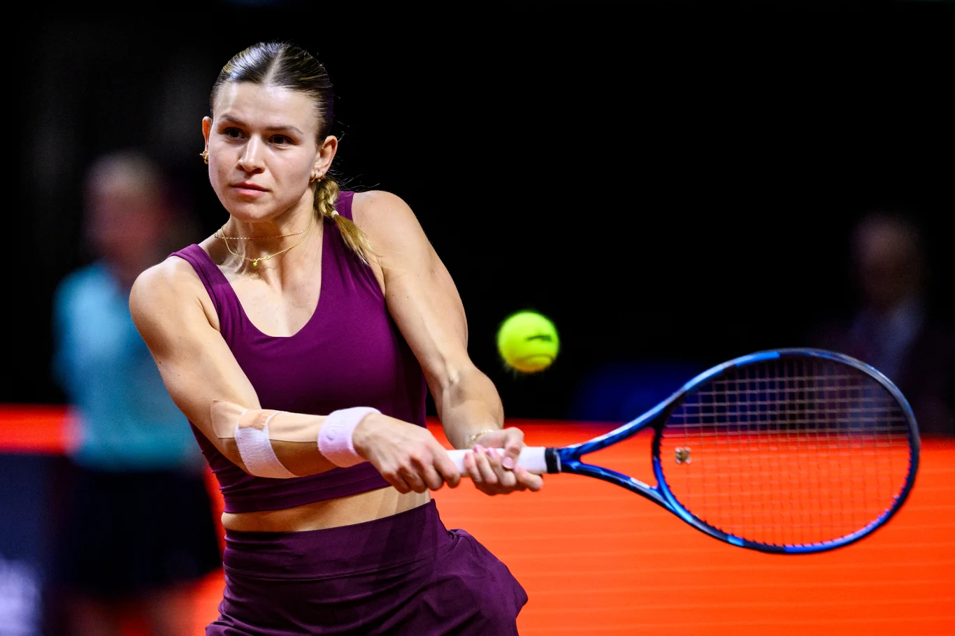 Kobieta w sportowym stroju uderza piłkę tenisową rakietą na korcie podczas meczu, skupiona i w ruchu, tło rozmazane z widocznym innym uczestnikiem.