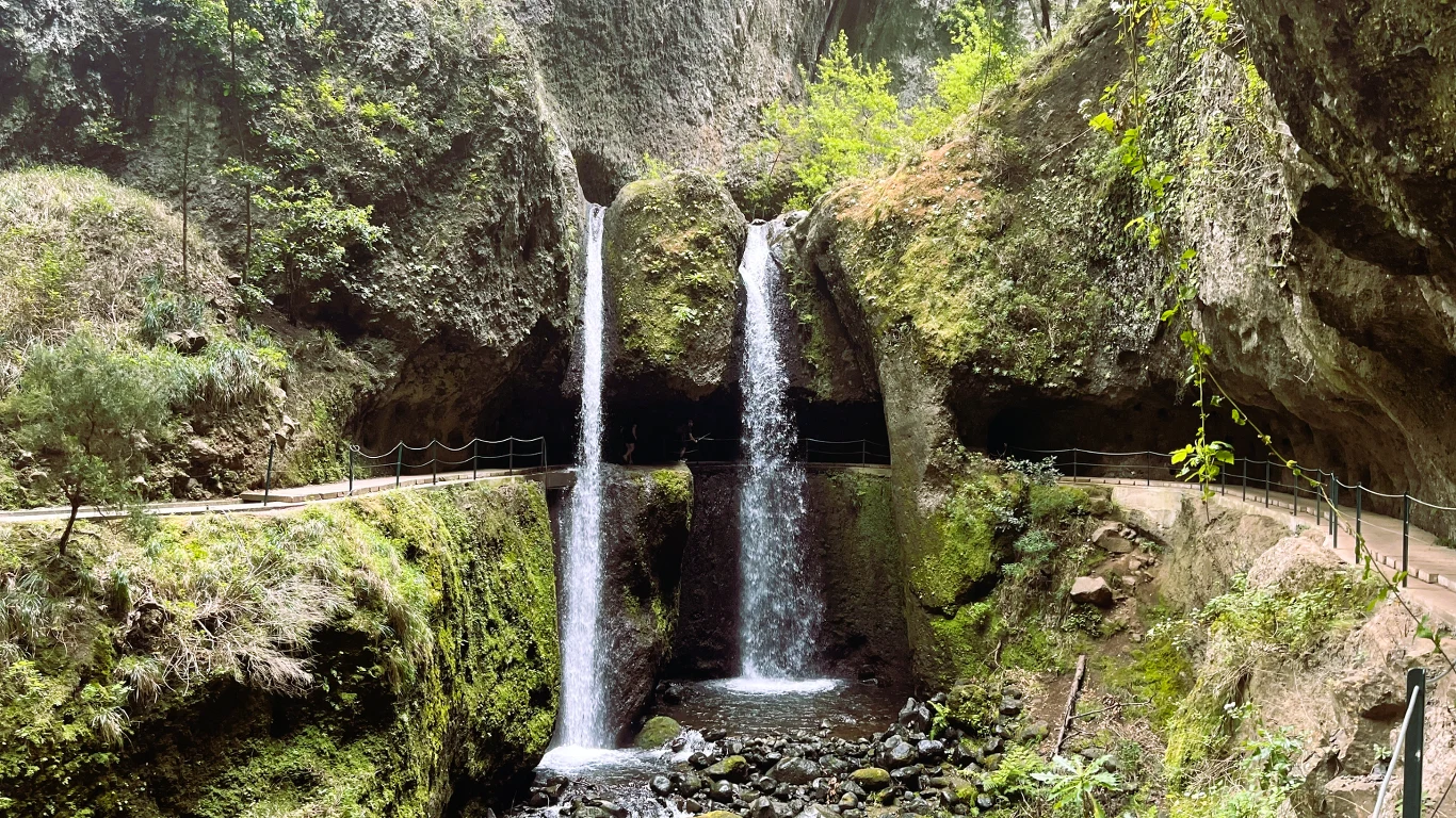 Dwa wodospady spływające z wysokiej skały do skalistego koryta rzeki, otoczone bujną zielenią oraz ścieżką zabezpieczoną barierkami, prowadzącą przez las i wokół wodospadu.