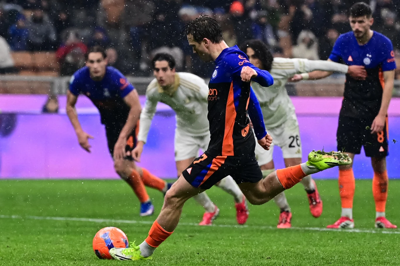 Piłkarz w trakcie wykonywania rzutu karnego na boisku, w tle kilku zawodników drużyny przeciwnej obserwujących moment strzału, widoczna intensywna koncentracja zawodnika oraz atmosfera stadionowa.