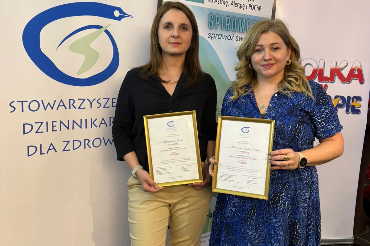 Dwie kobiety stoją obok siebie trzymając w rękach dyplomy, w tle widoczne są logotypy i banery związane z dziennikarskim stowarzyszeniem zdrowotnym.
