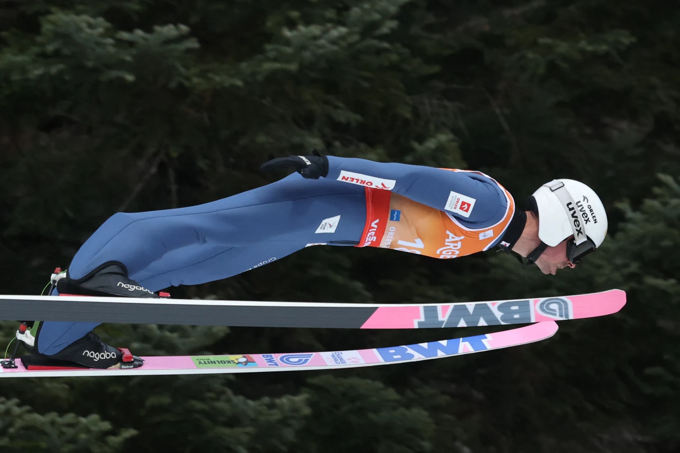 Skoczek narciarski w stroju sportowym podczas lotu, unosi się nad zeskokiem w klasycznej pozycji aerodynamicznej, tło stanowią drzewa pokryte igłami.