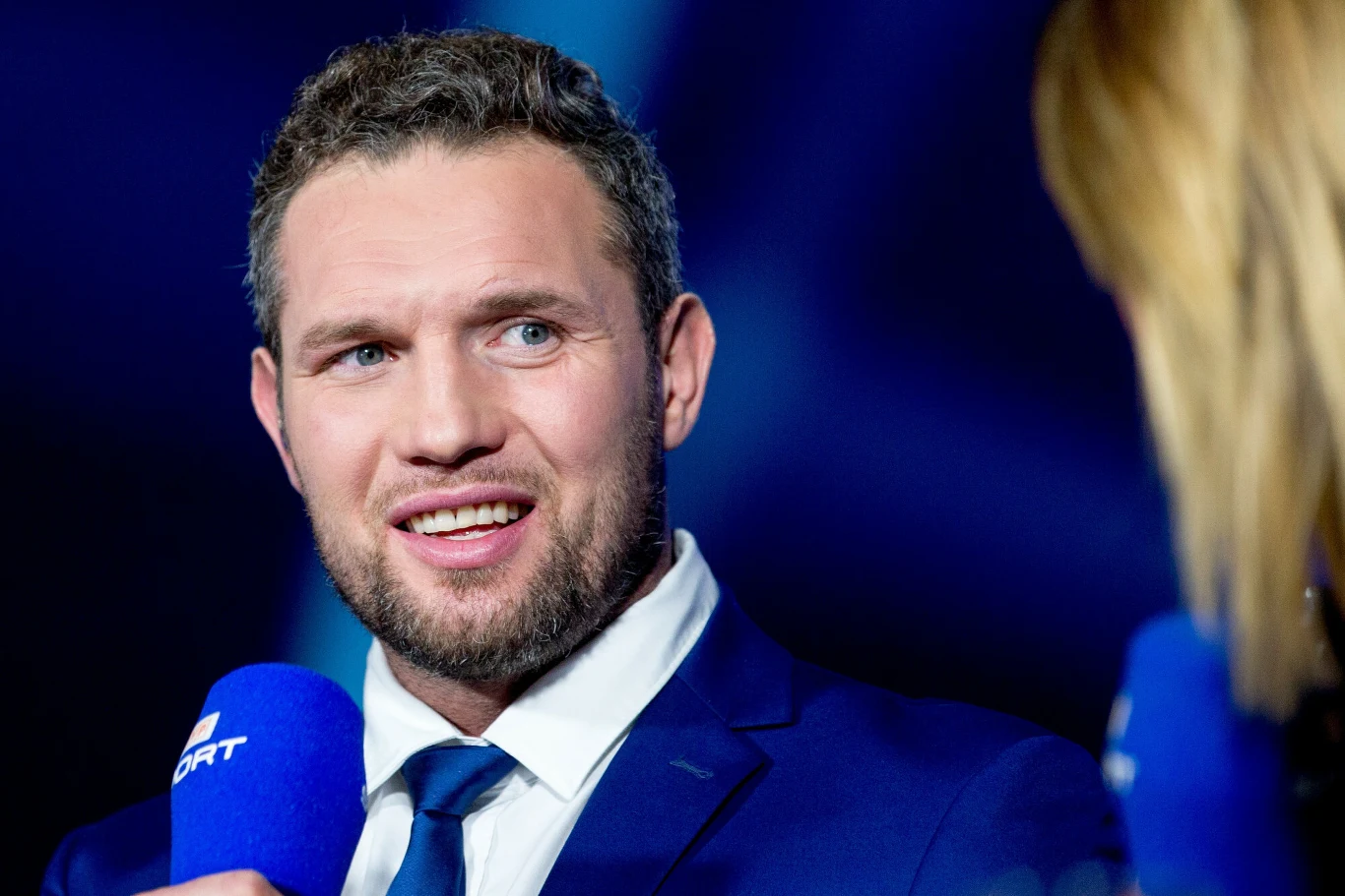 Mężczyzna w garniturze udziela wywiadu, trzyma w dłoni niebieski mikrofon z logo stacji sportowej, w tle rozmyte postacie i niebieskie światło.