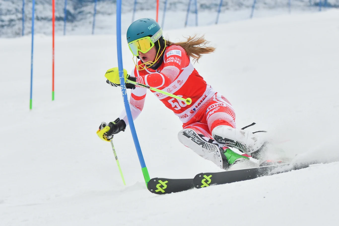 Aniela Sawicka Narciarka w intensywnym skręcie podczas slalomu, ubraną w czerwono-biały strój startowy z numerem 56, kask i gogle, przejeżdża pomiędzy niebieskimi i zielonymi tyczkami na zaśnieżonym stoku.