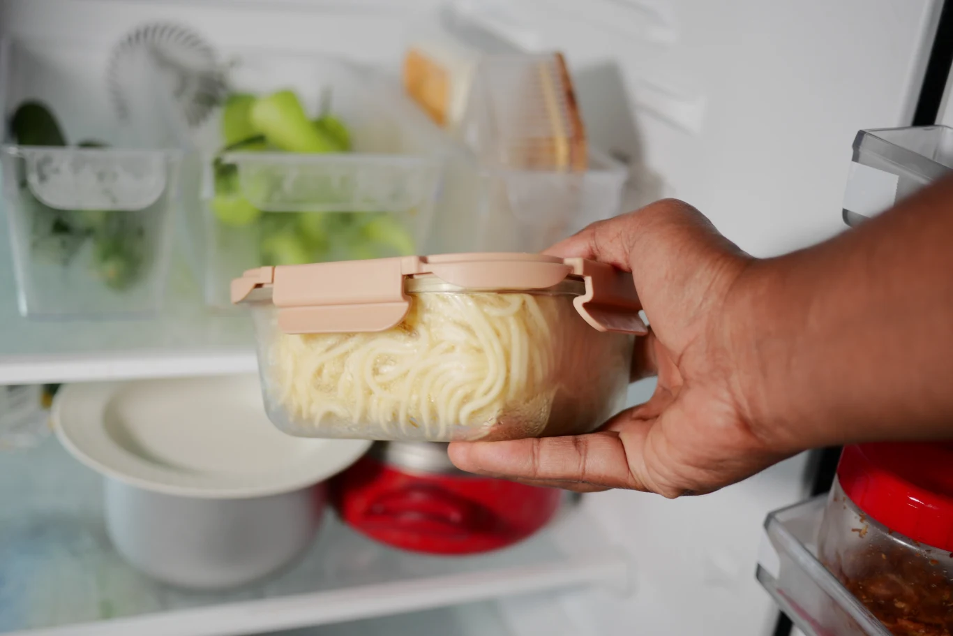 Ręka trzyma pojemnik z ugotowanym makaronem typu spaghetti, wkładanym do lodówki. W tle znajdują się przegródki z warzywami oraz inne produkty spożywcze, wszystkie ułożone na półkach chłodziarki.