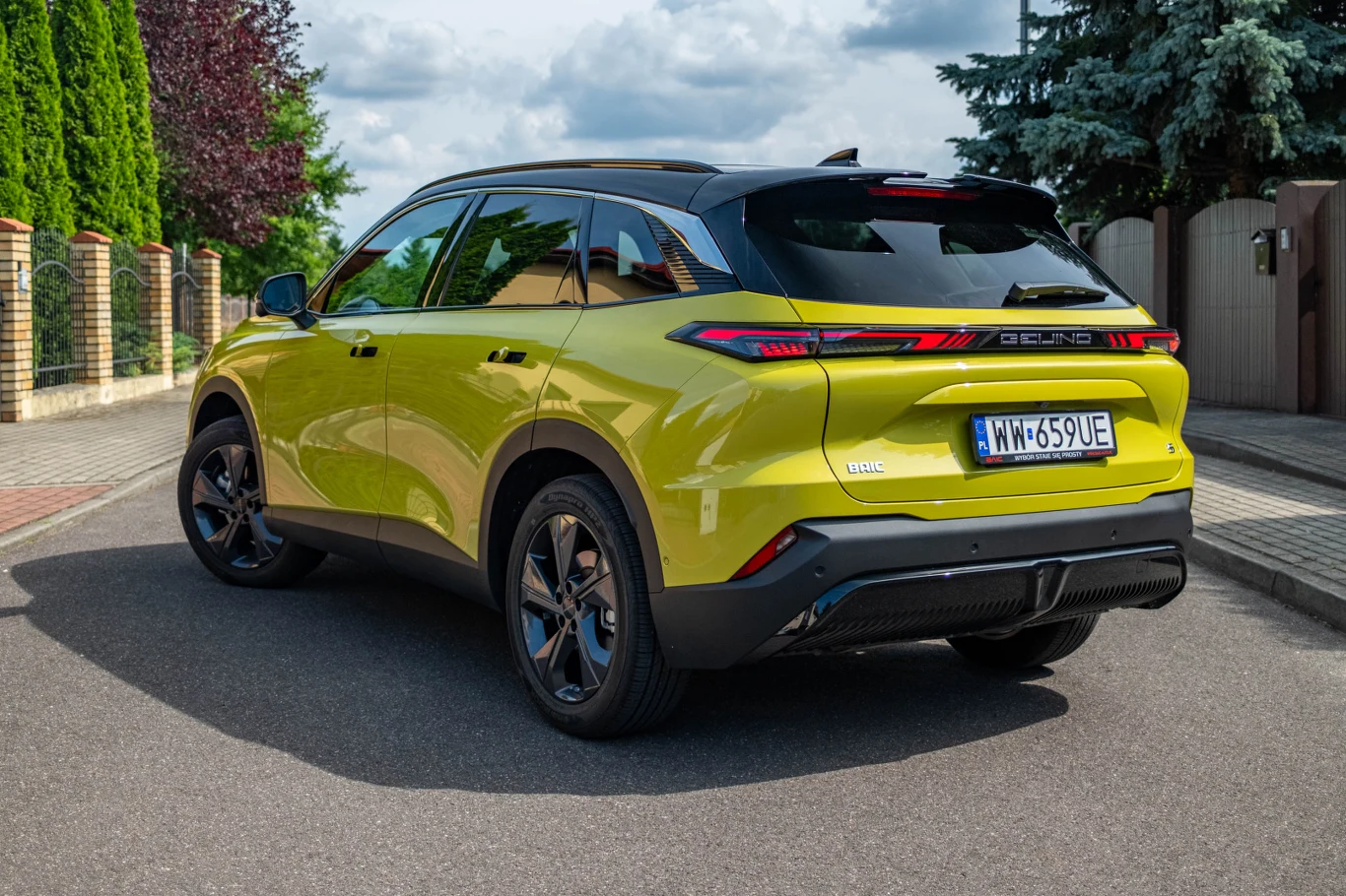 Jasnożółty samochód osobowy typu SUV marki Seres z czarnym dachem i nowoczesnym designem, zaparkowany na ulicy przy osiedlowym ogrodzeniu i otoczeniu zieleni.