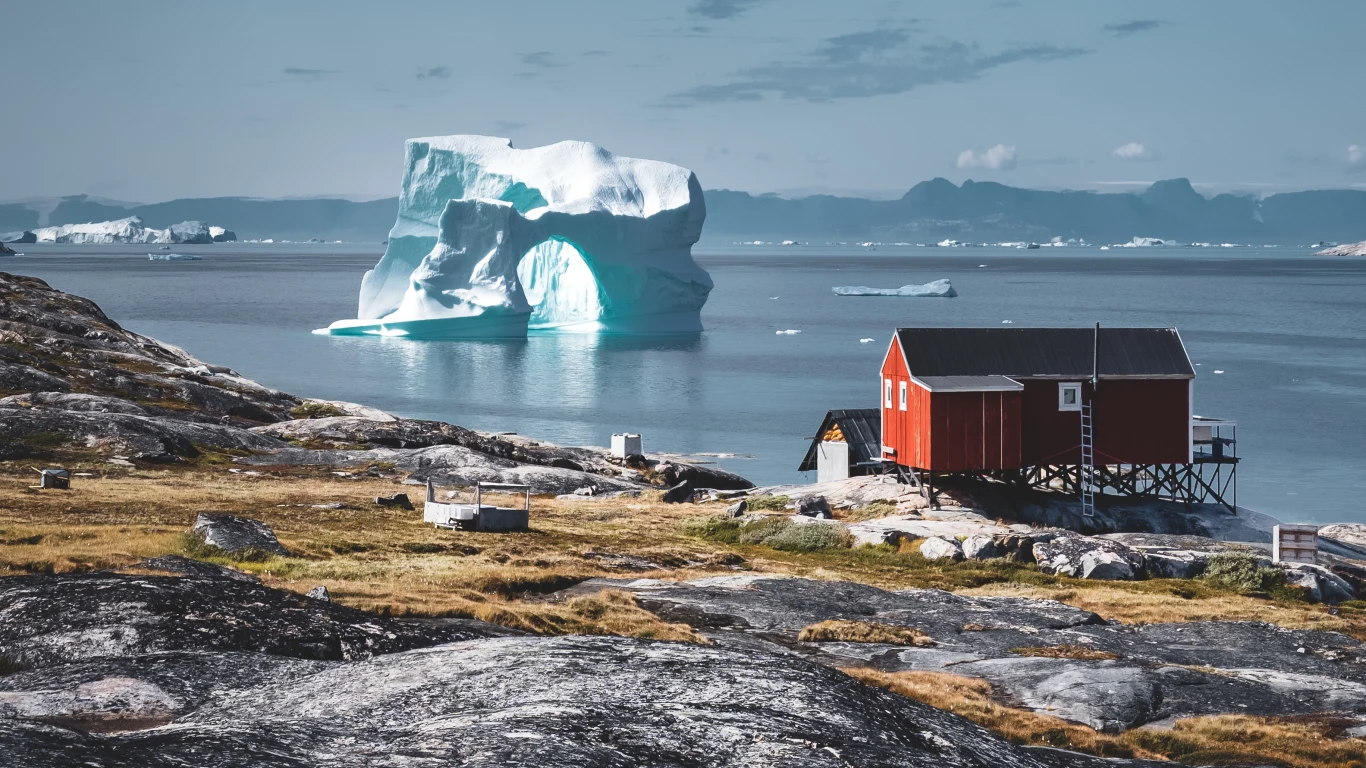 Ogromna biała góra lodowa unosząca się na spokojnych wodach fiordu, na pierwszym planie czerwona drewniana chata na palach, otoczona kamienistym i porośniętym mchem wybrzeżem.