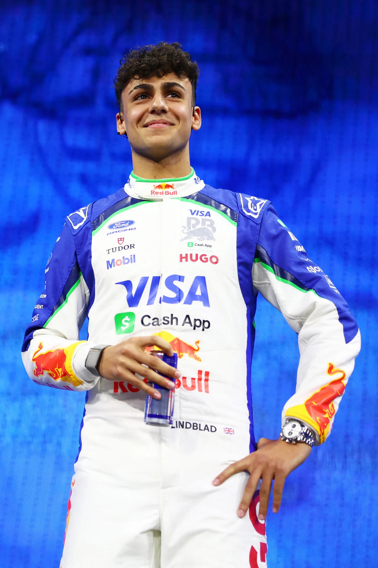 Młody mężczyzna w kombinezonie wyścigowym, opatrzonym licznymi logotypami sponsorów, trzyma puszkę napoju i stoi na tle niebieskiego ekranu. Wyraz zadowolenia na jego twarzy oraz profesjonalny strój wskazują na udział w wydarzeniu sportowym związanym z...