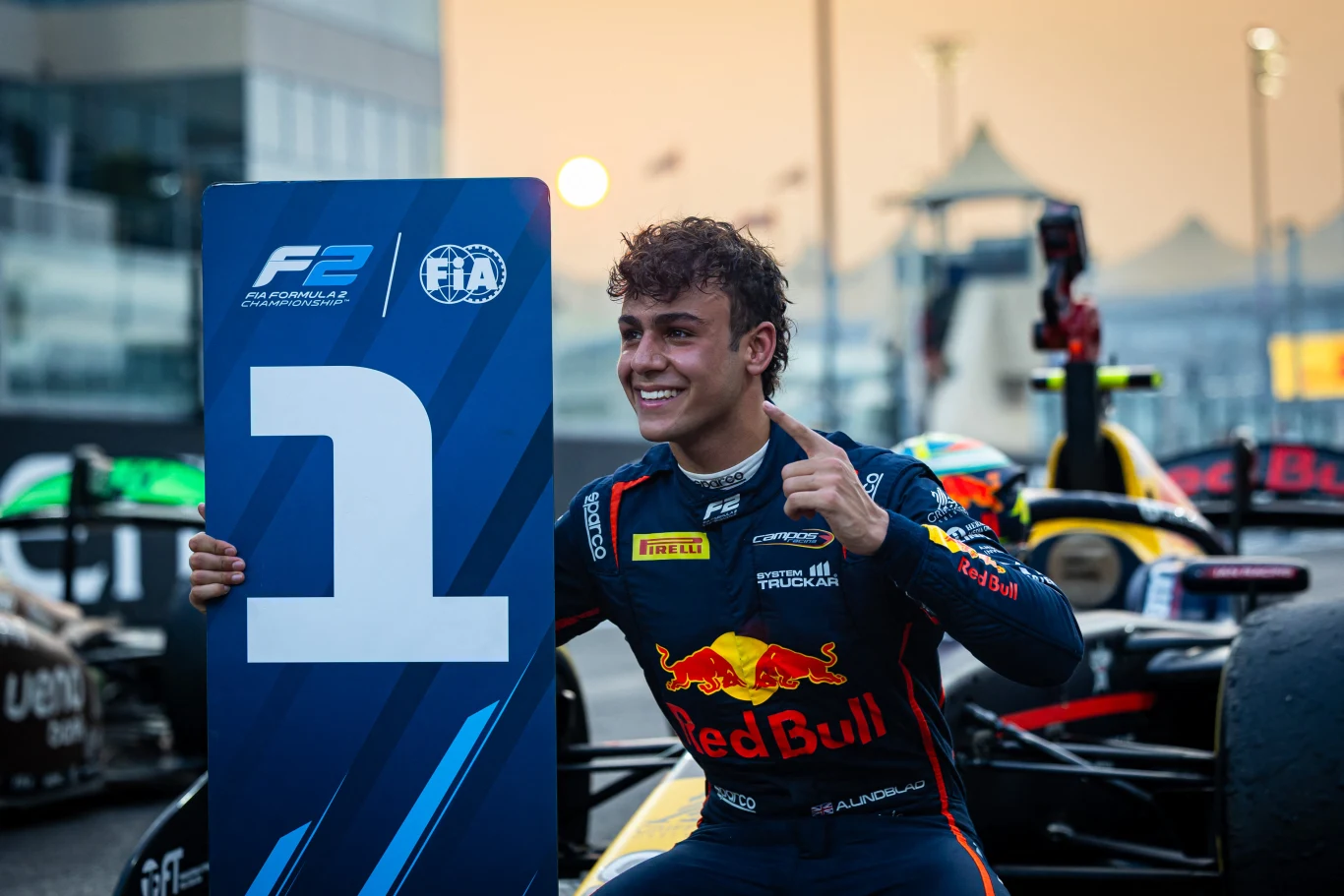 Młody kierowca wyścigowy w kombinezonie zespołu Red Bull siedzi obok tablicy z numerem jeden, wyraża radość pokazując gest zwycięstwa, w tle widoczne bolidy wyścigowe na torze.