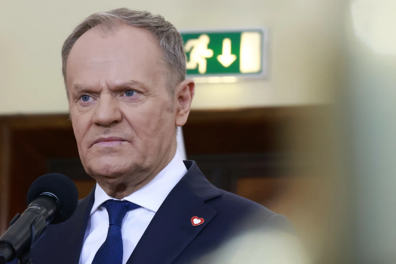 Premier Donald Tusk poinformował, że zamknięto schronisko w Sobolewie Mężczyzna w garniturze i krawacie stoi przed mikrofonem na tle jasnej ściany z widocznym znakiem ewakuacyjnym. Ubranie zdobi przypięta przypinka w kształcie czerwonego serca.