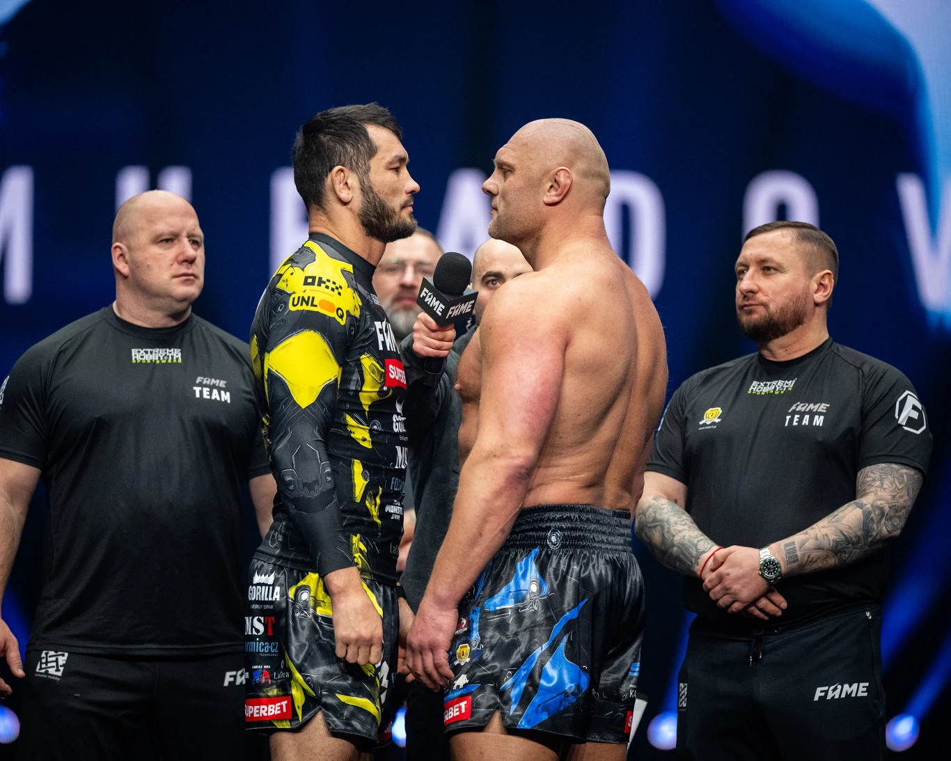 Dwóch umięśnionych mężczyzn stoi naprzeciwko siebie podczas oficjalnego spotkania sportowego, między nimi mikrofon, w tle stoją ochroniarze i członkowie zespołu, scena podświetlona reflektorami, atmosfera napięcia.