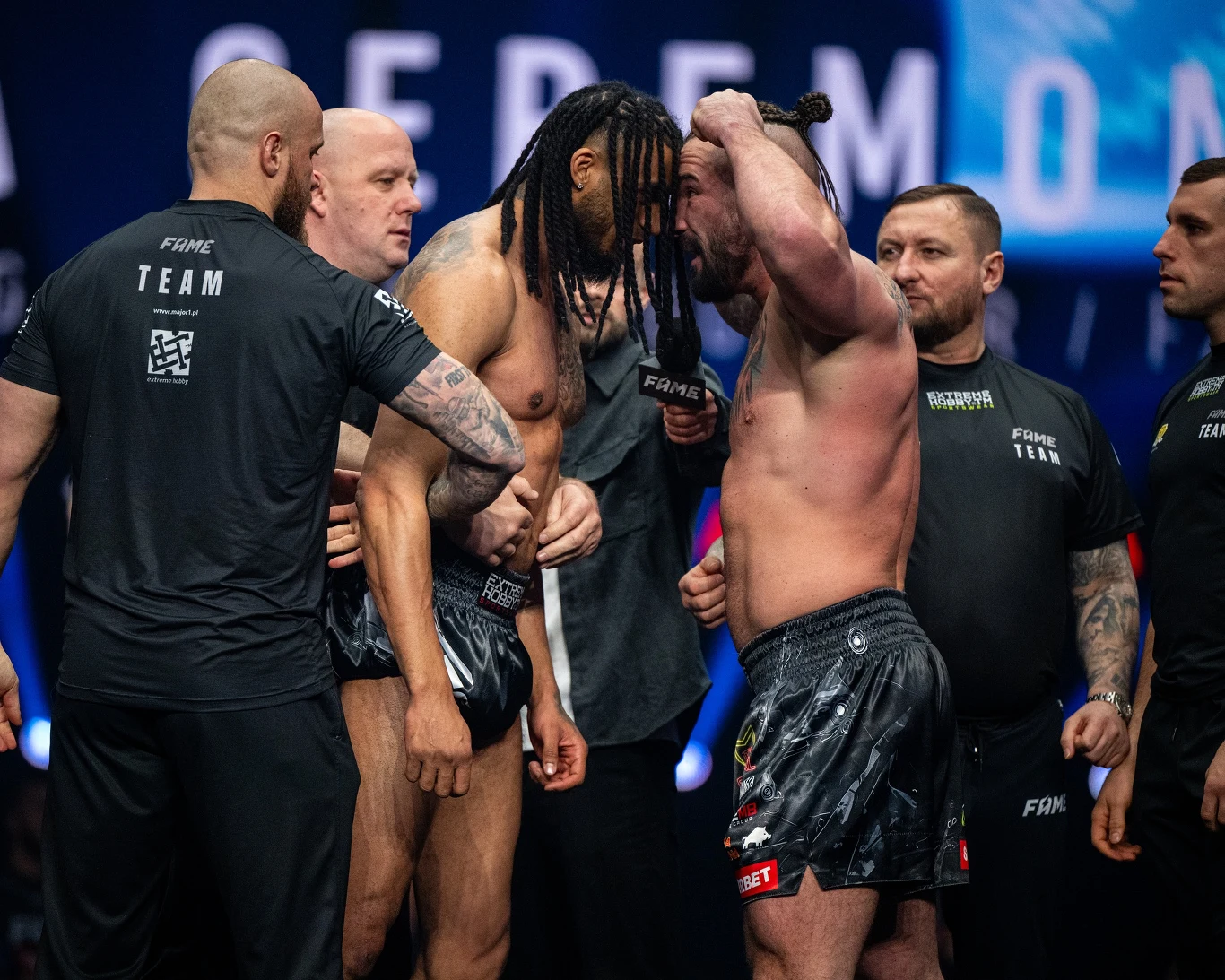 Dwóch umięśnionych mężczyzn w szortach sportowych stoi naprzeciwko siebie ze ściśle stykającymi się czołami, tworząc napiętą atmosferę rywalizacji, wokół nich kilku ochroniarzy i członków teamów bacznie obserwuje sytuację, a scena podkreśla charakter w...