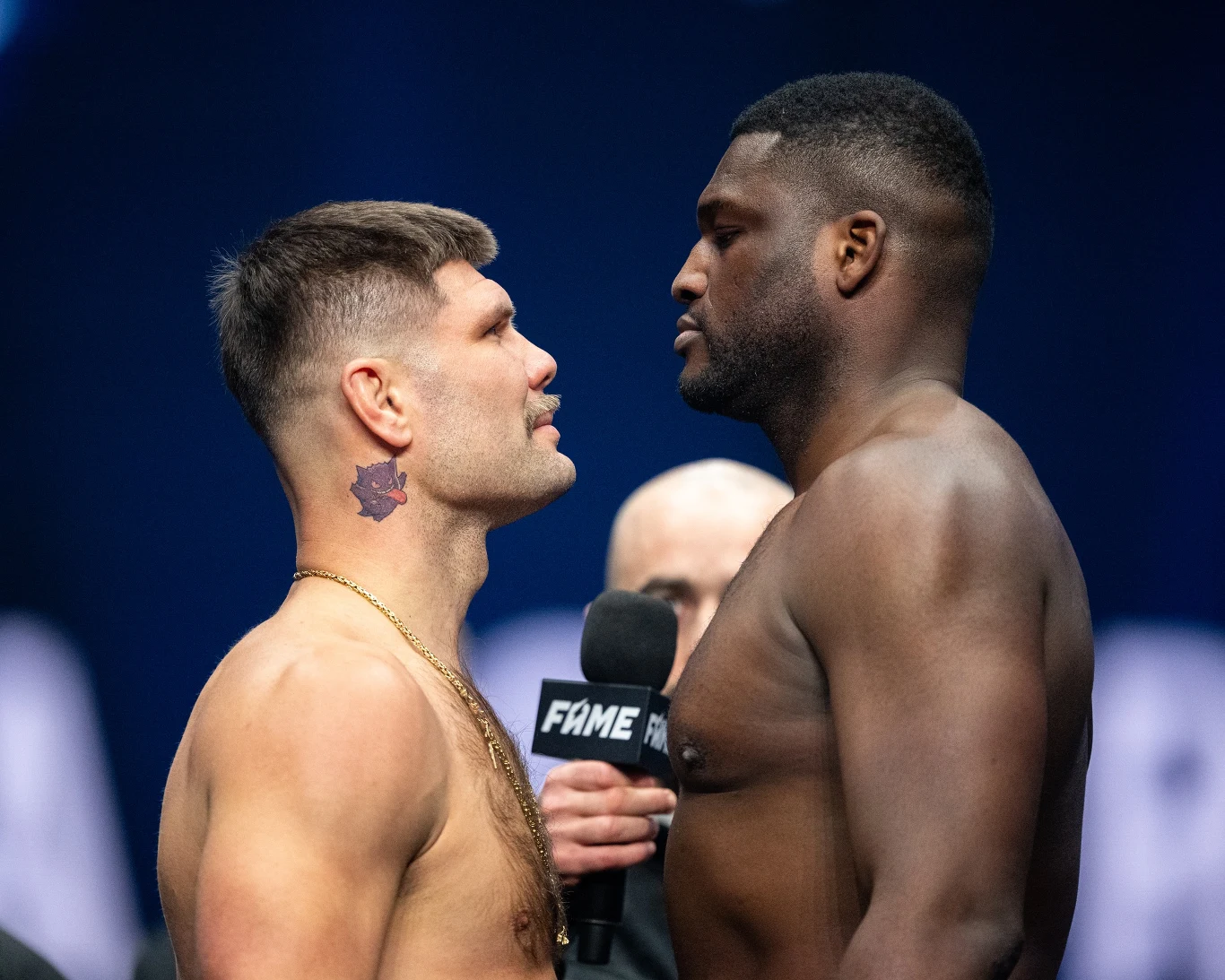 Dwóch umięśnionych mężczyzn stoi naprzeciwko siebie, patrząc sobie prosto w oczy podczas oficjalnego spotkania przed walką. Jeden z nich ma widoczny tatuaż na szyi, w tle osoba z mikrofonem i logo organizacji sportowej.