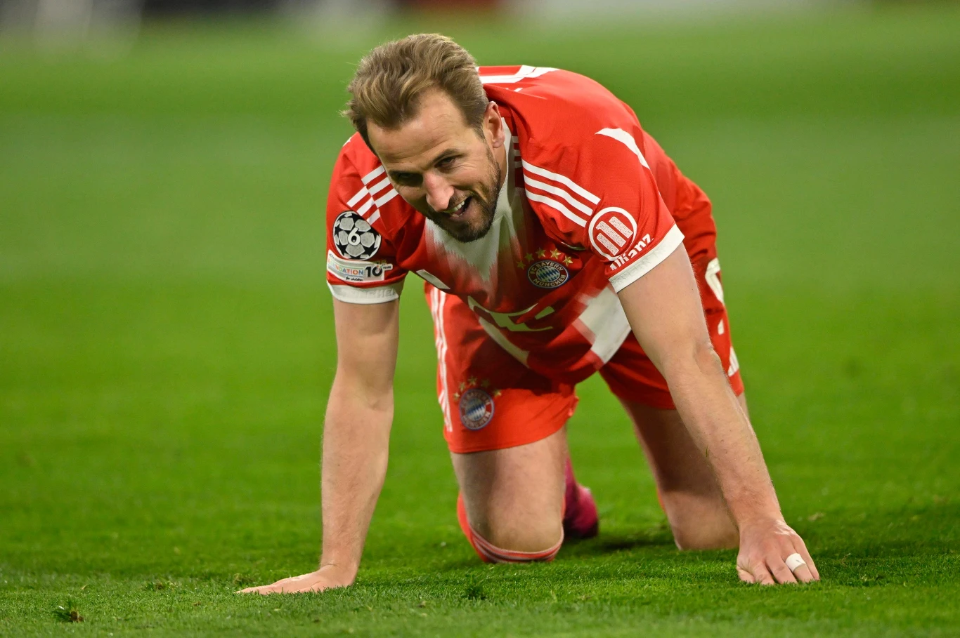 Harry Kane Piłkarz w czerwono-białym stroju drużyny Bayernu Monachium znajduje się na murawie w pozycji na kolanach i wspiera się rękami o ziemię, wyrażając zmęczenie lub chwilowe osłabienie podczas meczu.
