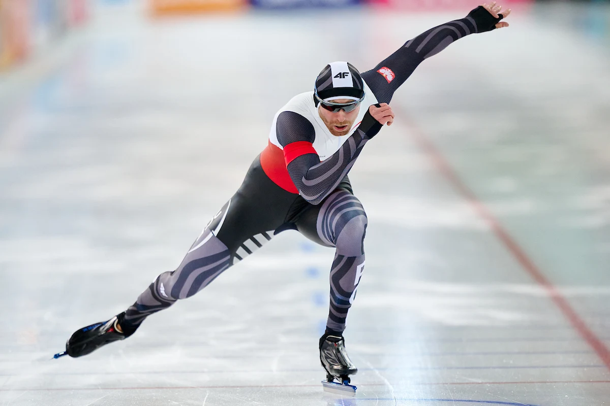 Damian Żurek Łyżwiarz szybki w profesjonalnym stroju podczas wyścigu na lodowisku, dynamiczna pozycja z jedną ręką wyciągniętą do przodu i drugą do tyłu, widoczne oznaczenia sportowe na stroju.