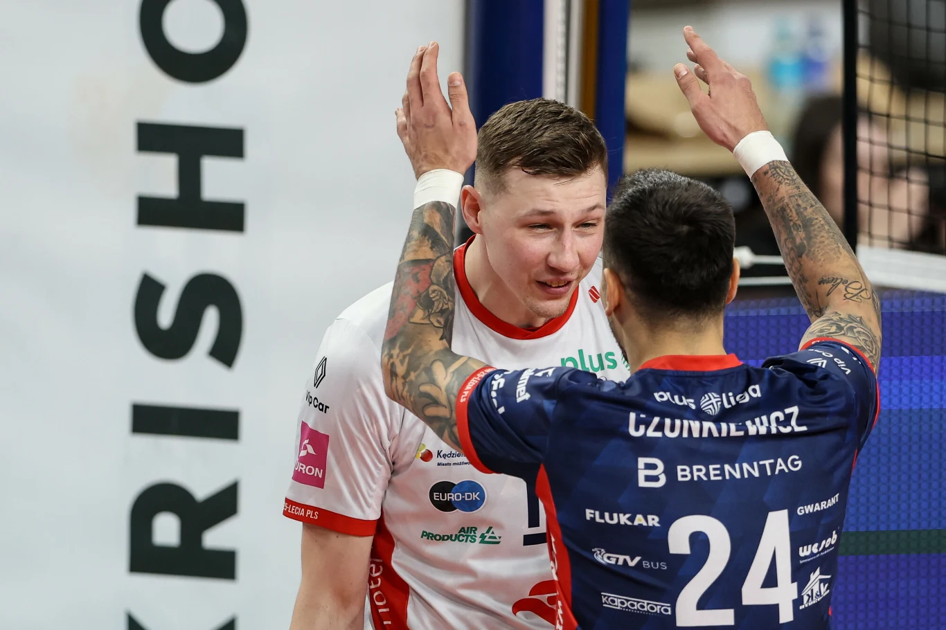 Dwóch siatkarzy podczas meczu, jeden w białej koszulce, drugi w granatowej, stoją bardzo blisko siebie, podnosząc ręce, prawdopodobnie wyrażając emocje po zakończonej akcji.