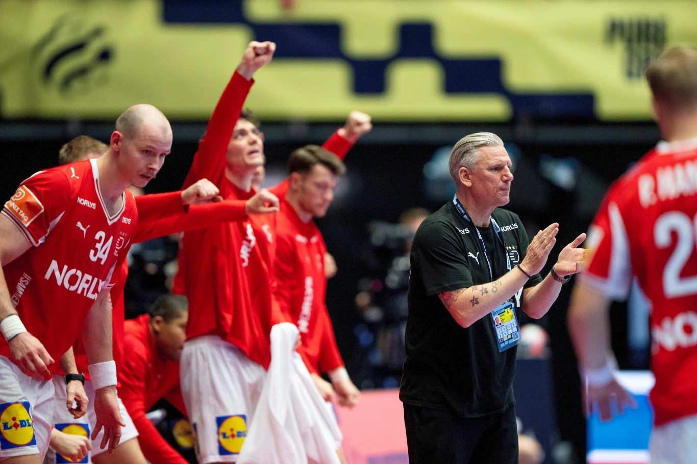 Trener Nikolaj Jacobsen i reprezentanci Danii Zawodnicy drużyny piłki ręcznej w czerwonych strojach oraz ich trener stoją na linii bocznej boiska, okazując silne emocje i zaangażowanie, niektórzy mają wzniesione pięści w geście triumfu, trener bije brawo.