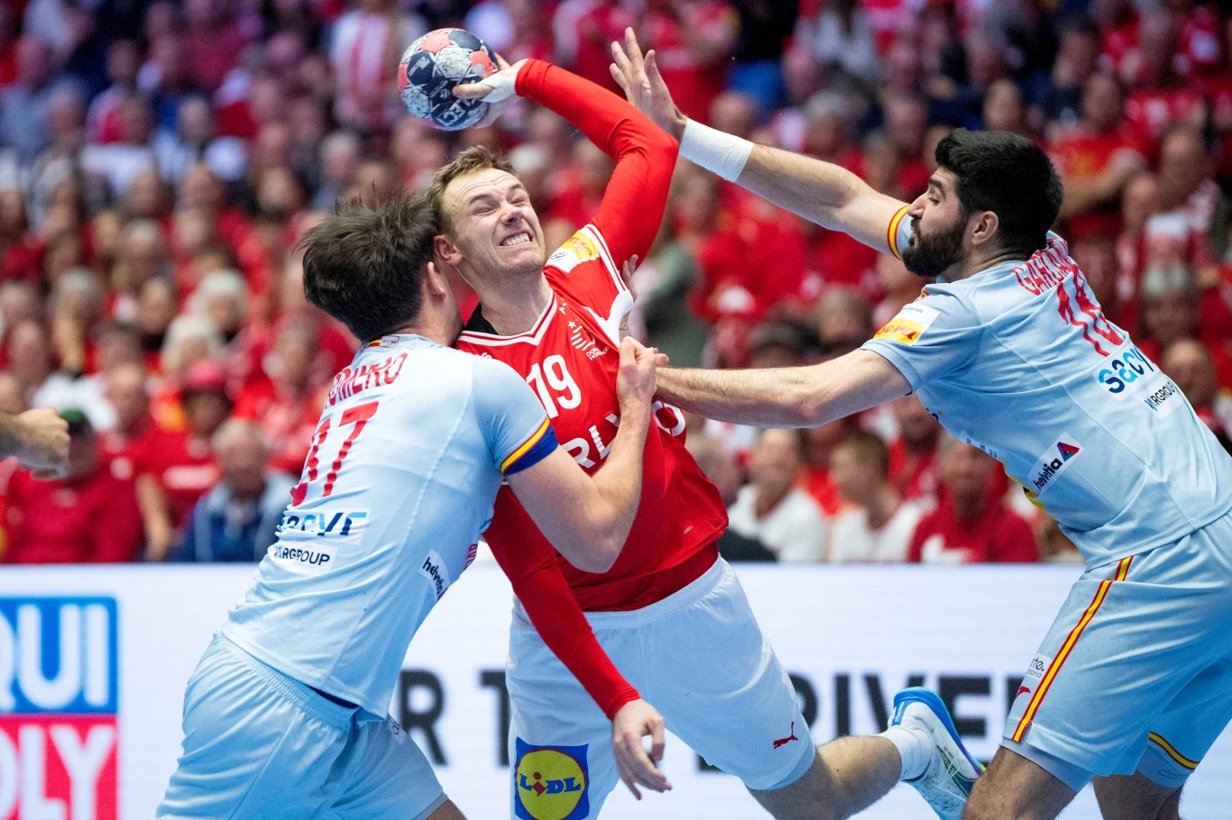 Mathias Gidsel zatrzymywany przez Hiszpanów Dynamiczna akcja meczowa piłki ręcznej, zawodnik w czerwonym stroju próbuje rzucić piłkę, mocno naciskany przez dwóch przeciwników w białych strojach, tłum kibiców w tle.