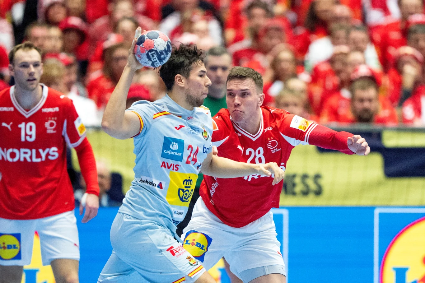 Daniel Dujszebajew w meczu z Danią Zawodnik w jasnym stroju próbuje rzucić piłkę ręczną podczas meczu, będąc bronionym przez przeciwnika w czerwonym stroju. W tle widoczna grupa kibiców ubrana na czerwono oraz pozostali zawodnicy.