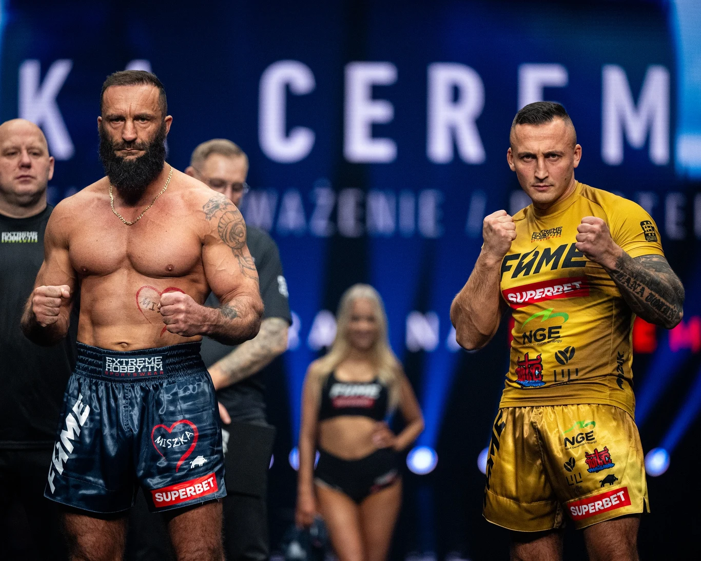 Dwóch umięśnionych zawodników MMA pozuje do zdjęcia w ringu, jeden ubrany w czarne spodenki, drugi w złote, obaj z uniesionymi pięściami. W tle widoczni sędziowie i hostessy oraz światła sceniczne.