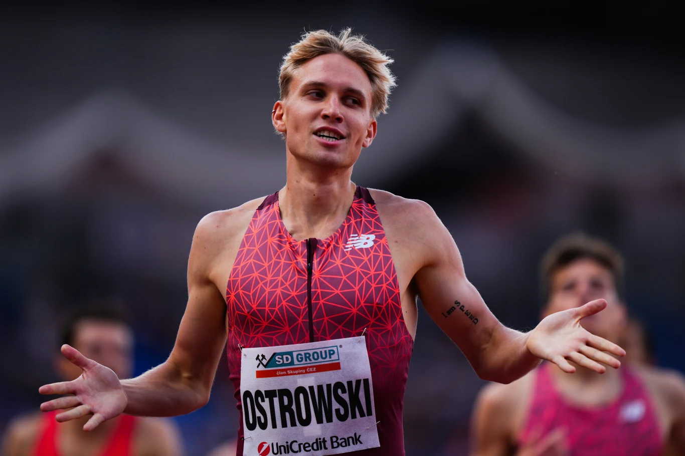 Lekkoatleta w stroju sportowym gestykuluje rozkładając ręce, na piersi ma numer startowy z nazwiskiem Ostrowski. W tle widoczni zamazani zawodnicy i zarys stadionu.