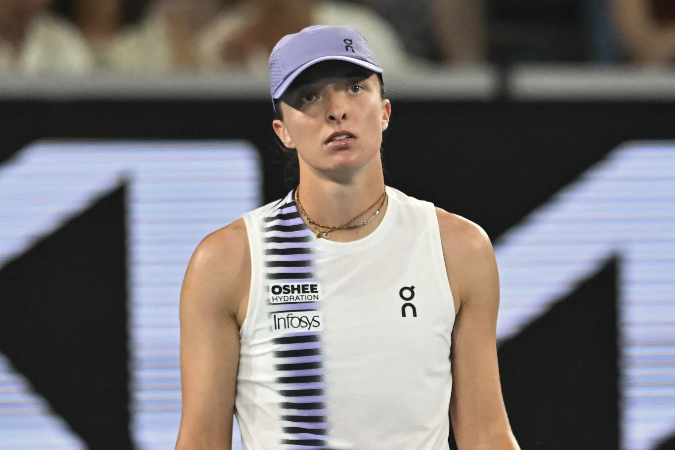 Tenisistka w sportowej koszulce i fioletowej czapce z daszkiem stoi na korcie, patrząc przed siebie z poważnym wyrazem twarzy, w tle rozmyta publiczność.