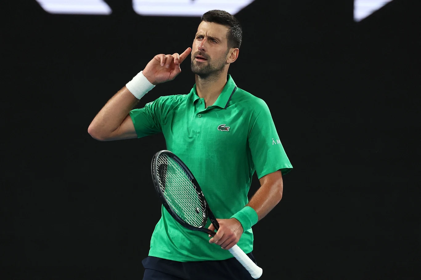 Tenisista w zielonej koszulce wykonuje gest palcem przy uchu, trzymając rakietę tenisową na korcie.