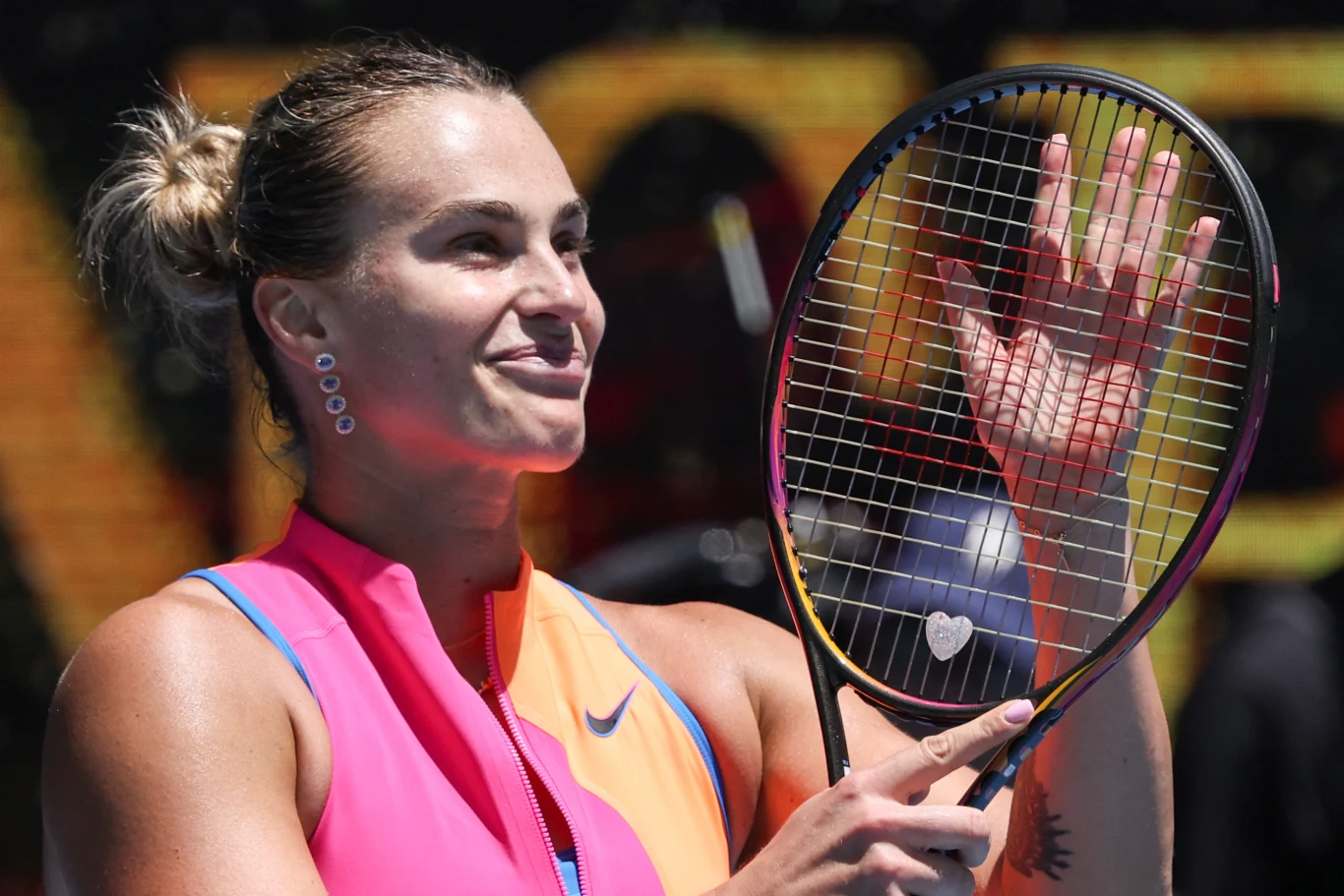 Kobieta ubrana w sportowy strój tenisowy trzyma rakietę, uśmiecha się i unosi otwartą dłoń w geście pozdrowienia podczas meczu tenisowego.