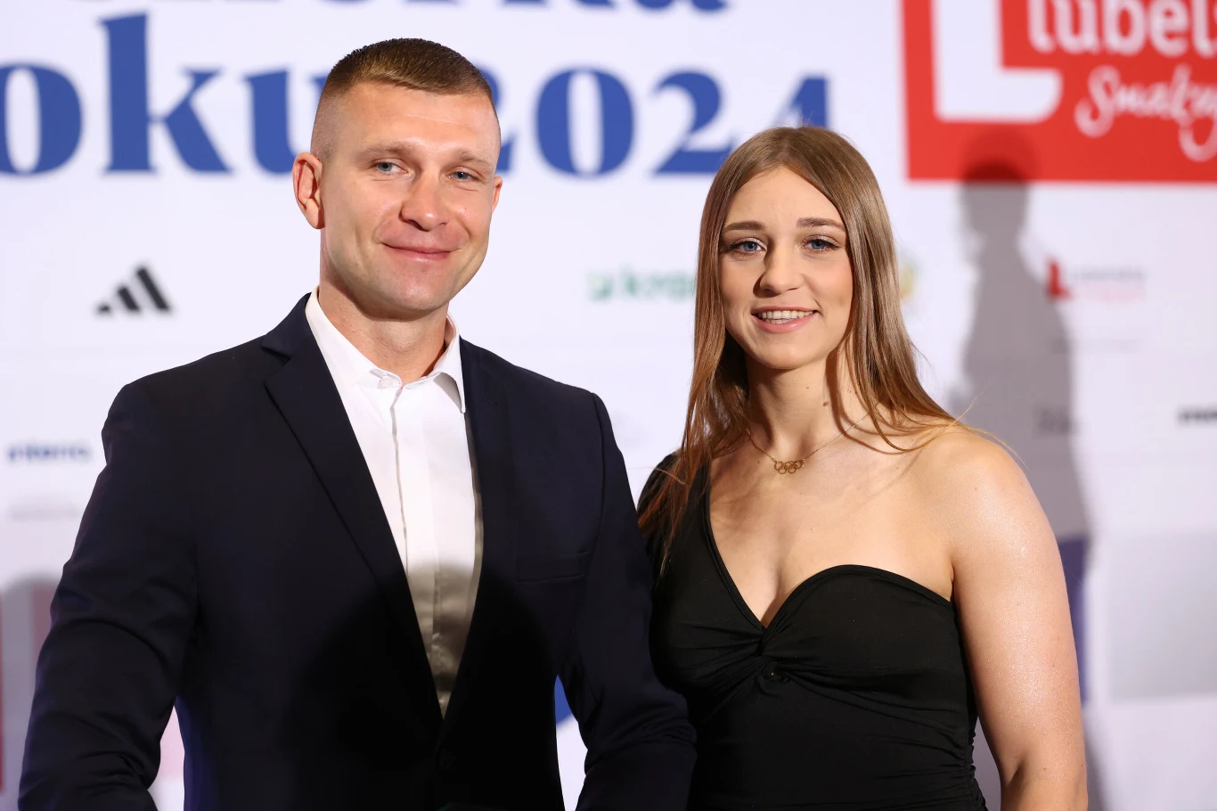 Mężczyzna w ciemnym garniturze i jasnej koszuli stoi obok kobiety w czarnej sukience na tle ścianki z logotypami i napisem '2024'. Oboje uśmiechają się, sprawiają wrażenie uczestników oficjalnego wydarzenia.