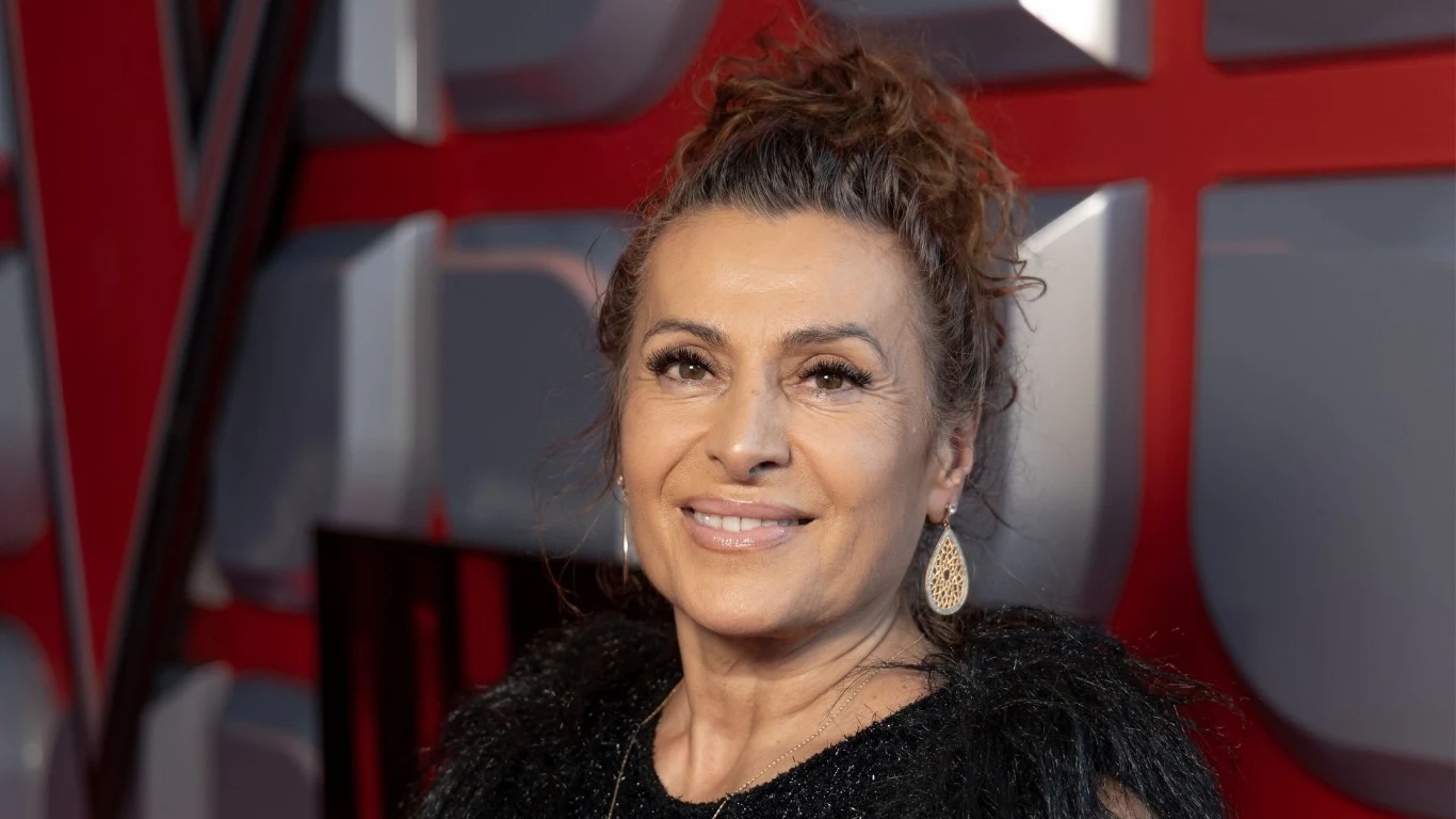 Violetta Kapcewicz wywołała gorące emocje w "The Voice Senior" Uśmiechnięta kobieta z upiętymi włosami i dużymi kolczykami wisi na tle czerwono-szarej dekoracji scenicznej.