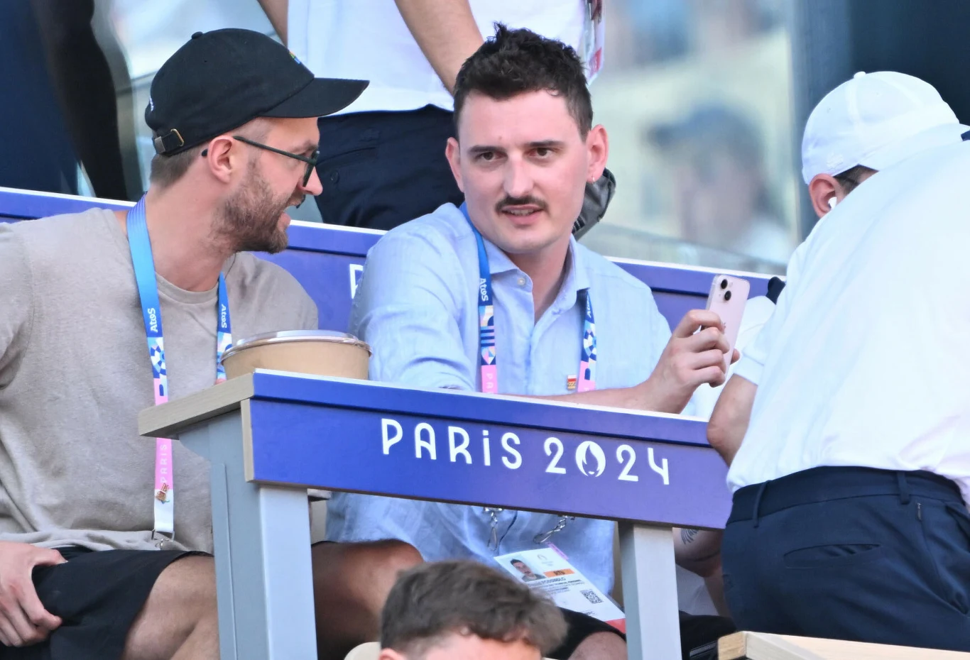 Dawid Podsiadło na aukcję WOŚP wystawił możliwość występu z nim na PGE Narodowym Dwóch mężczyzn siedzi na trybunach oznaczonych logo Paris 2024, rozmawiają ze sobą, jeden z nich trzyma smartfona, obaj noszą identyfikatory kibiców lub uczestników imprezy.
