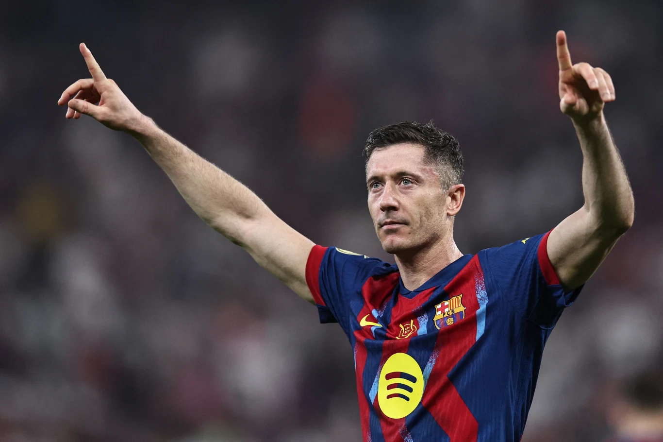 Robert Lewandowski Piłkarz w koszulce FC Barcelony z uniesionymi rękami celebruje sukces na boisku sportowym, w tle niewyraźna publiczność stadionu.