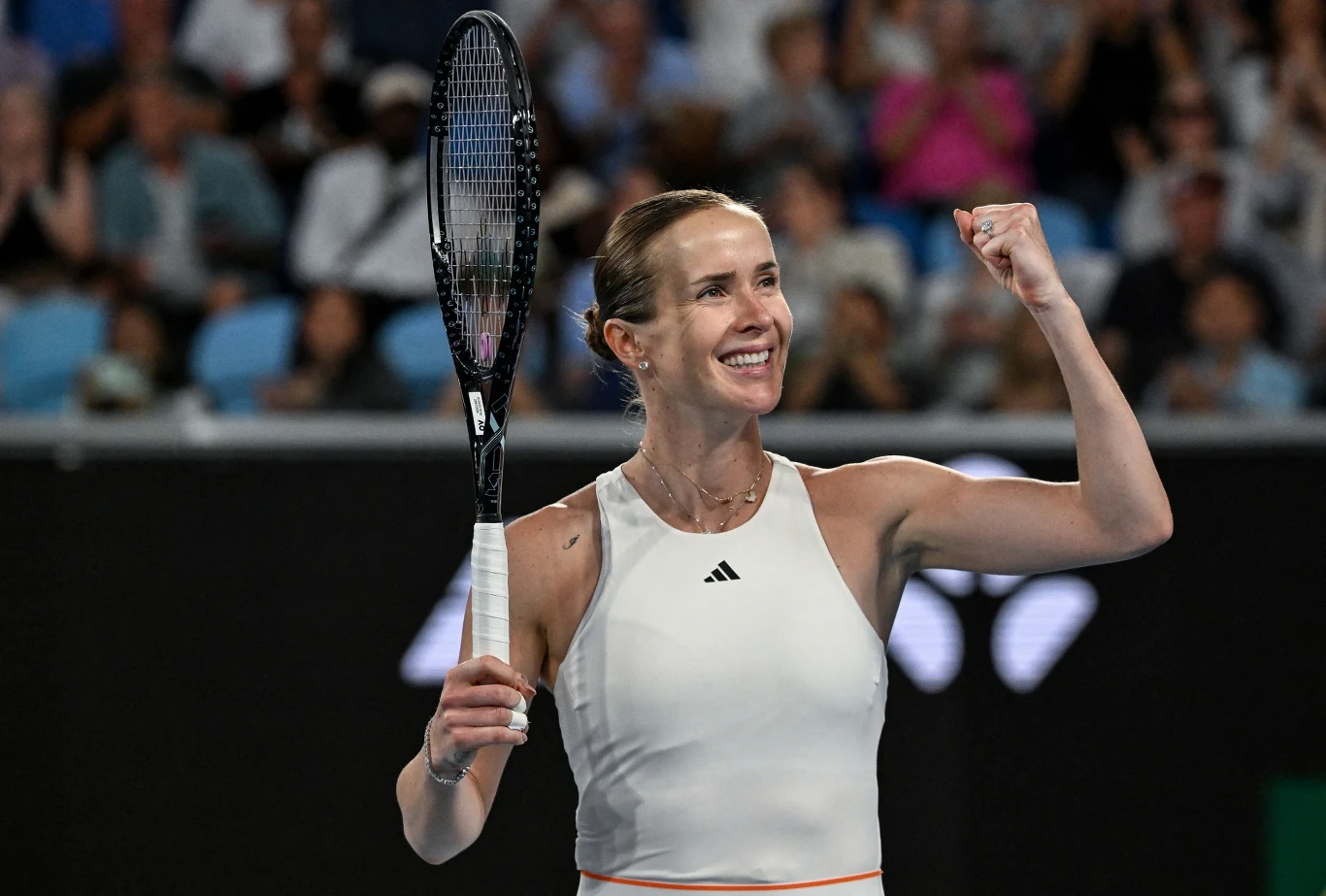 Tenisistka w białym stroju sportowym unosi zaciśniętą pięść i uśmiecha się, trzymając rakietę tenisową. W tle rozmyta publiczność na trybunach obserwująca wydarzenie.