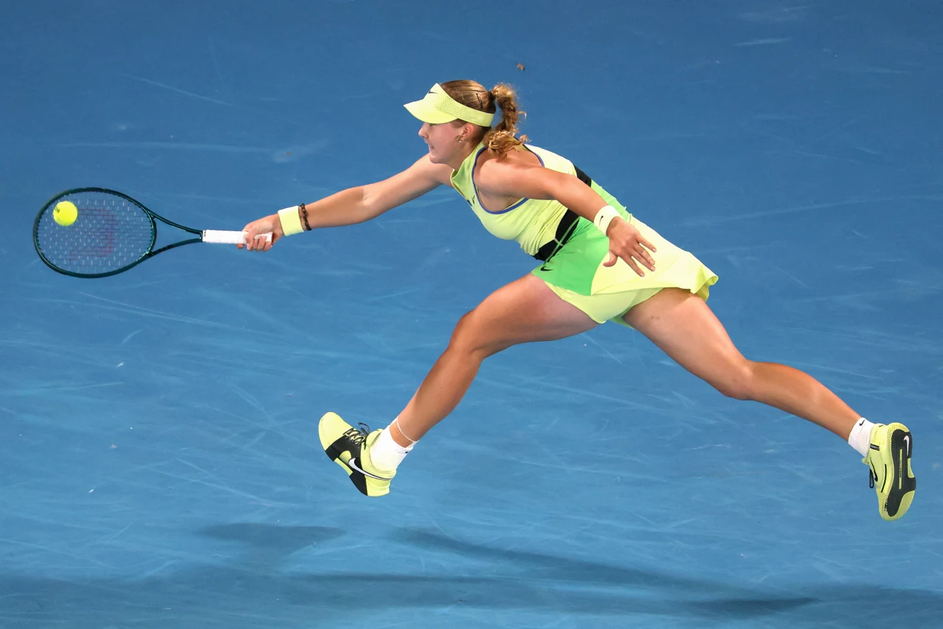 Tenisistka w żółtej sukience wykonuje dynamiczny wślizg, wyciągając rakietę, by odbić lecącą piłkę na niebieskim korcie.