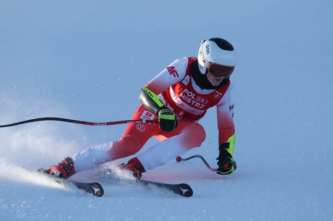 Narciarz w biało-czerwonym kombinezonie podczas dynamicznego skrętu na stoku, wyposażony w kask i gogle, z widocznym logo "Polski Mistrz" na stroju, otoczony błękitnym śniegiem.