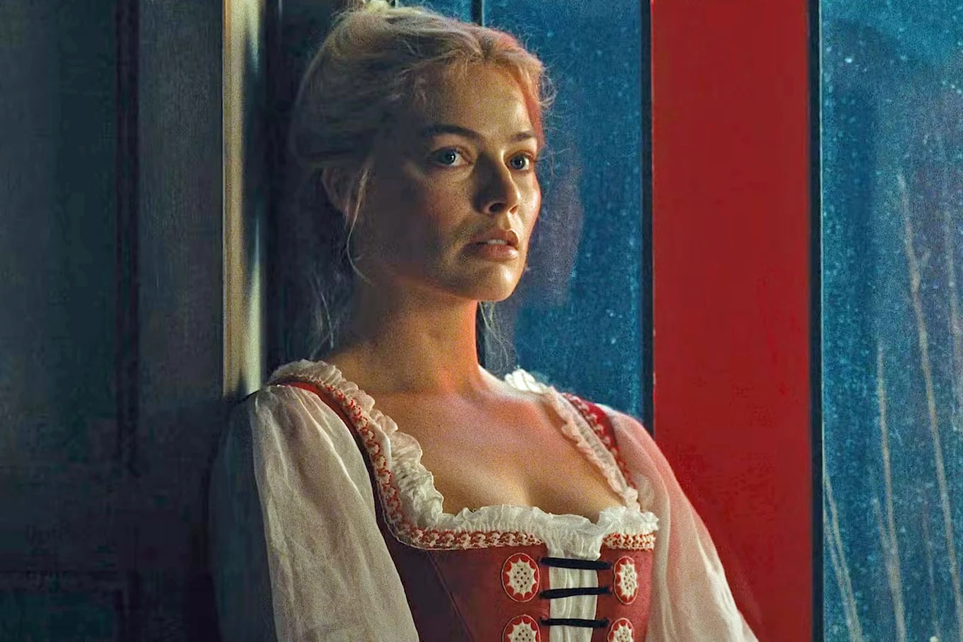 Margot Robbie w scenie z filmu "Wichrowe wzgórza" Kobieta o blond włosach ubrana w tradycyjny strój z białą bluzką i czerwonym gorsetem stoi oparta o drewnianą ścianę, za nią widoczne są czerwone okienne ramy.