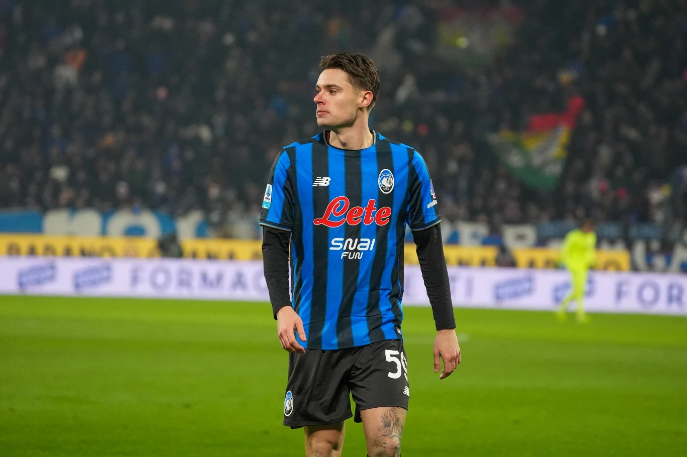 Piłkarz w stroju meczowym Atalanty Bergamo na tle stadionu, za nim widoczni kibice i rozmyte sylwetki innych uczestników wydarzenia sportowego.