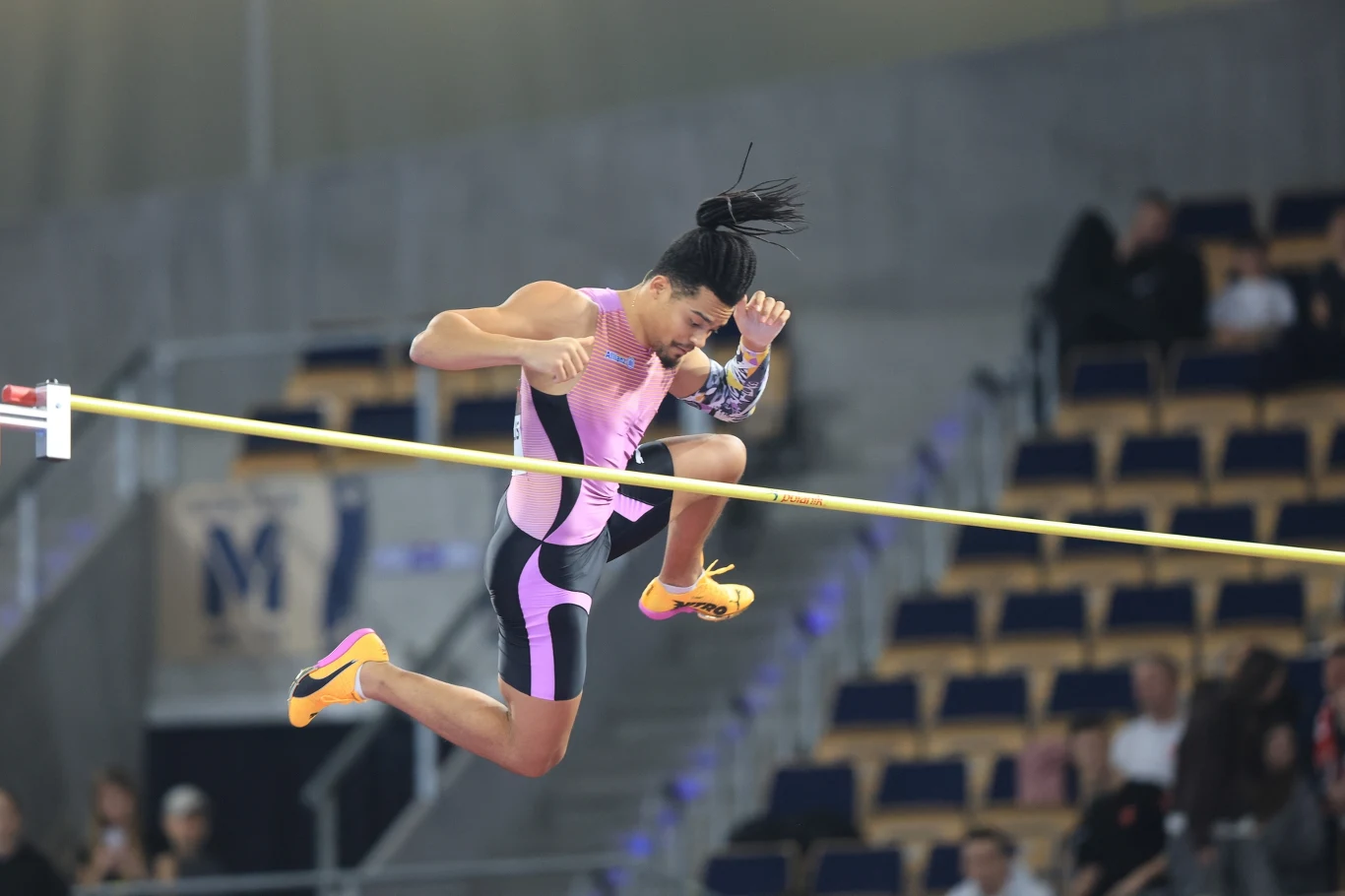 Lekkoatleta w różowym stroju wykonuje skok wzwyż podczas zawodów halowych, dynamicznie przekraczając poprzeczkę w pełnej koncentracji. W tle puste i częściowo zajęte trybuny.