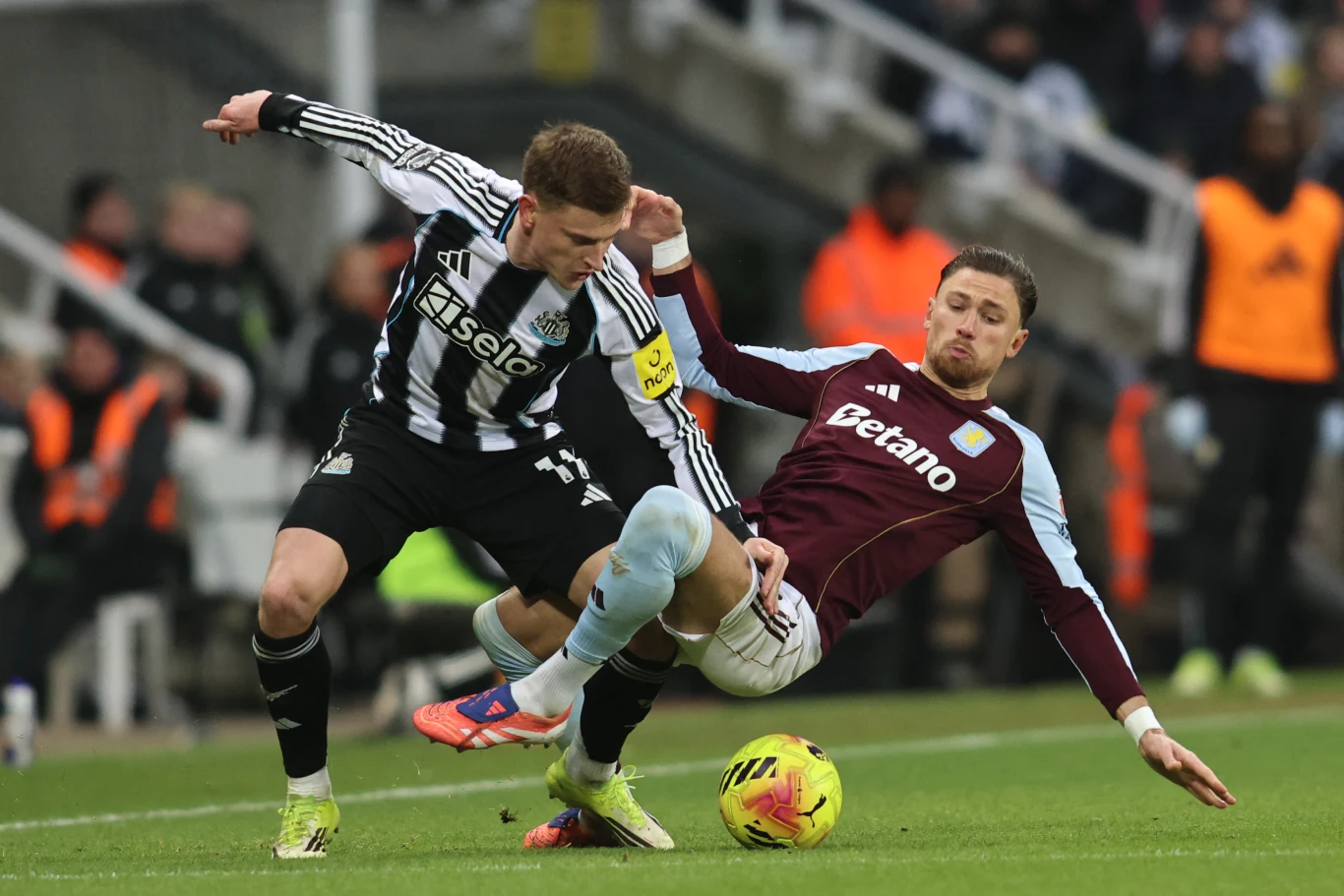 Dwóch piłkarzy rywalizuje o piłkę podczas meczu, jeden z nich wykonuje wślizg, starając się przejąć kontrolę nad futbolówką, w tle widoczna jest część trybun oraz inni uczestnicy spotkania.
