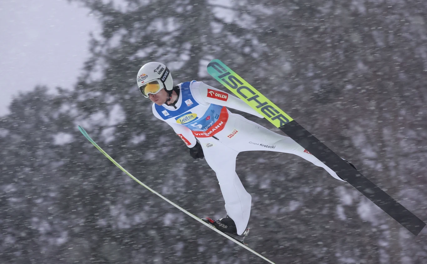 Skoczek narciarski podczas lotu na tle padającego śniegu, ubrany w biały kombinezon z widocznymi logotypami sponsorów i kaskiem, szeroko rozłożone narty, tło stanowią ciemne, zamglone drzewa.