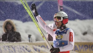 Skoczek narciarski w kombinezonie sportowym, z nartami w dłoni, stojący na śniegu, unosi rękę w geście pozdrowienia, w tle rozmyte postacie widzów i zaśnieżone trybuny.