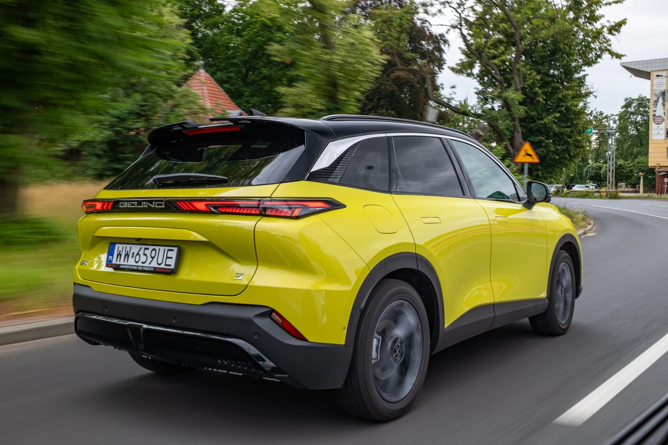 Żółty nowoczesny samochód typu SUV jadący po zakręcie drogi miejskiej, w otoczeniu zielonych drzew oraz budynków w tle, tablice rejestracyjne wskazujące na rejestrację pojazdu w Polsce.