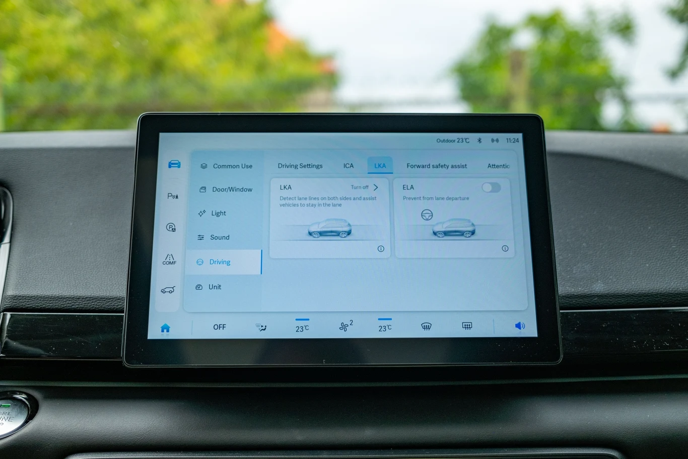 Ekran komputera pokładowego w samochodzie z wyświetlonymi ustawieniami jazdy oraz asystentami bezpieczeństwa. W tle przez przednią szybę widoczne rozmyte drzewa i fragment nieba.