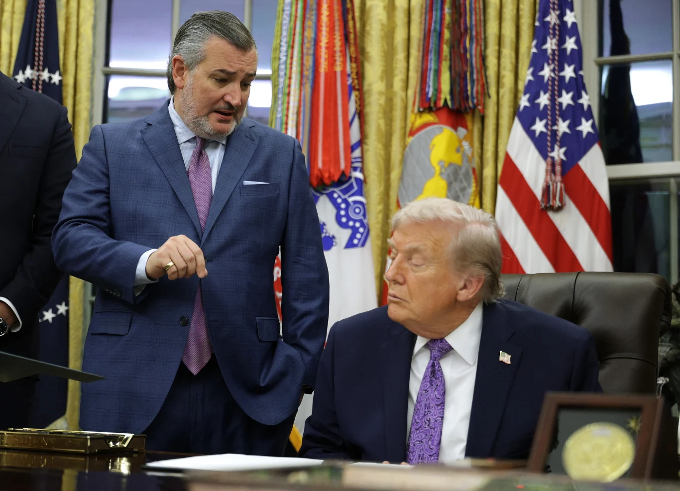 Senator Ted Cruz krytykuje politykę Donalda Trumpa Dwóch polityków w eleganckich garniturach podczas rozmowy w gabinecie, jeden z nich siedzi za biurkiem, a drugi gestykuluje stojąc obok. W tle znajdują się flagi amerykańskie i wojskowe, ciężkie zasłony oraz złote elementy dekoracyjne.