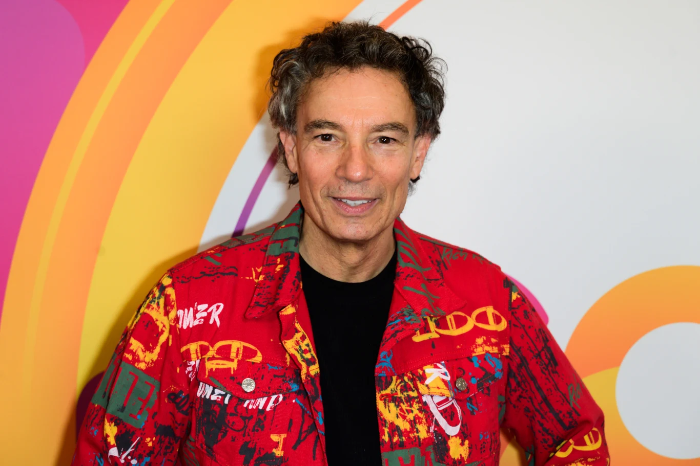 Jacek Stachursky obchodzi 60. urodziny Mężczyzna o siwych lokowanych włosach ubrany w kolorową, czerwoną koszulę z różnorodnymi nadrukami, stoi na tle kolorowej, abstrakcyjnej grafiki z okręgami i łukami w odcieniach żółci, pomarańczu i różu.