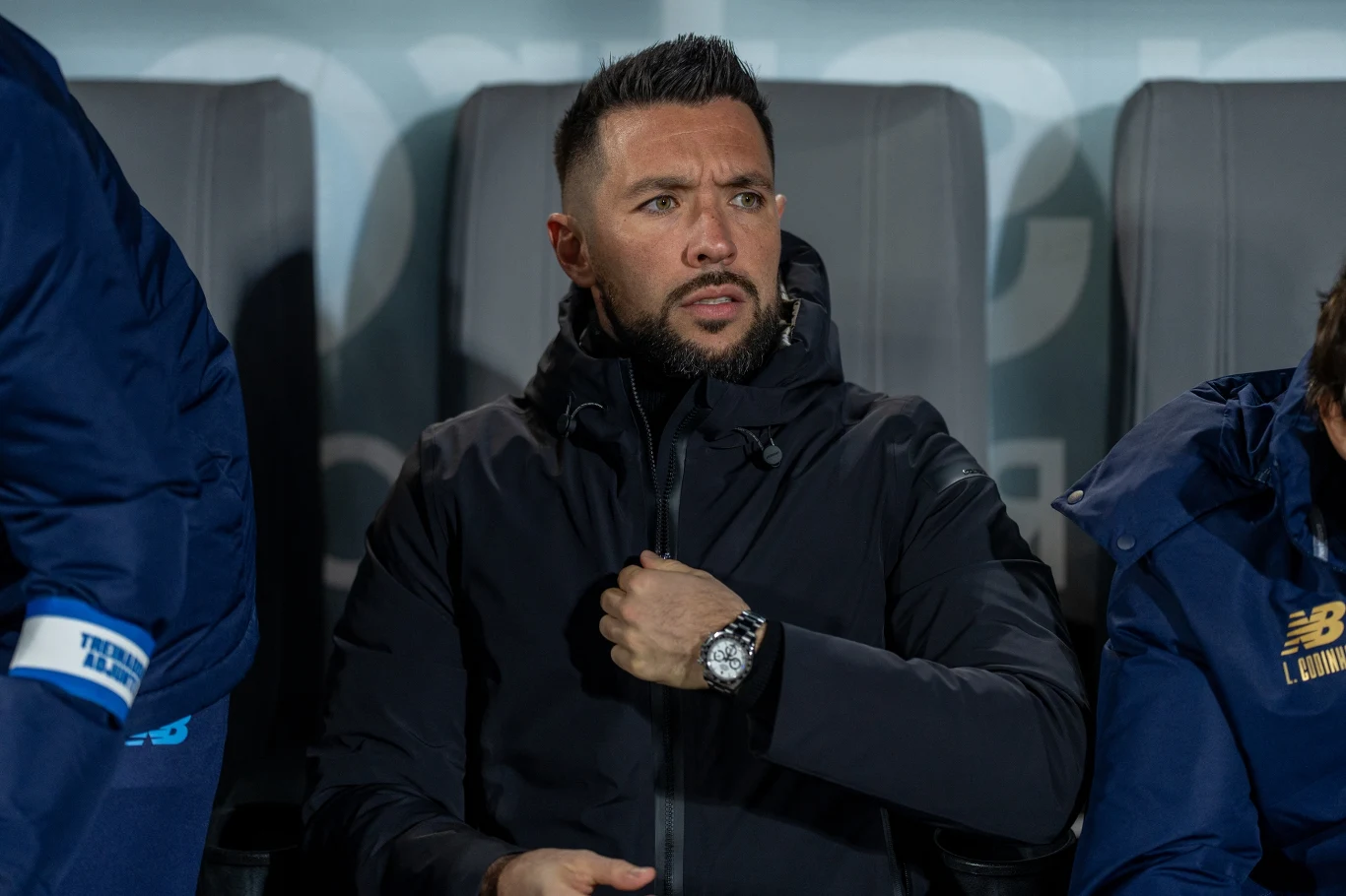 Mężczyzna w czarnej kurtce siedzi na stadionowej ławce rezerwowych, zapięty pod szyję, patrzy w bok z poważnym wyrazem twarzy, obok niego inne osoby w sportowych ubraniach.